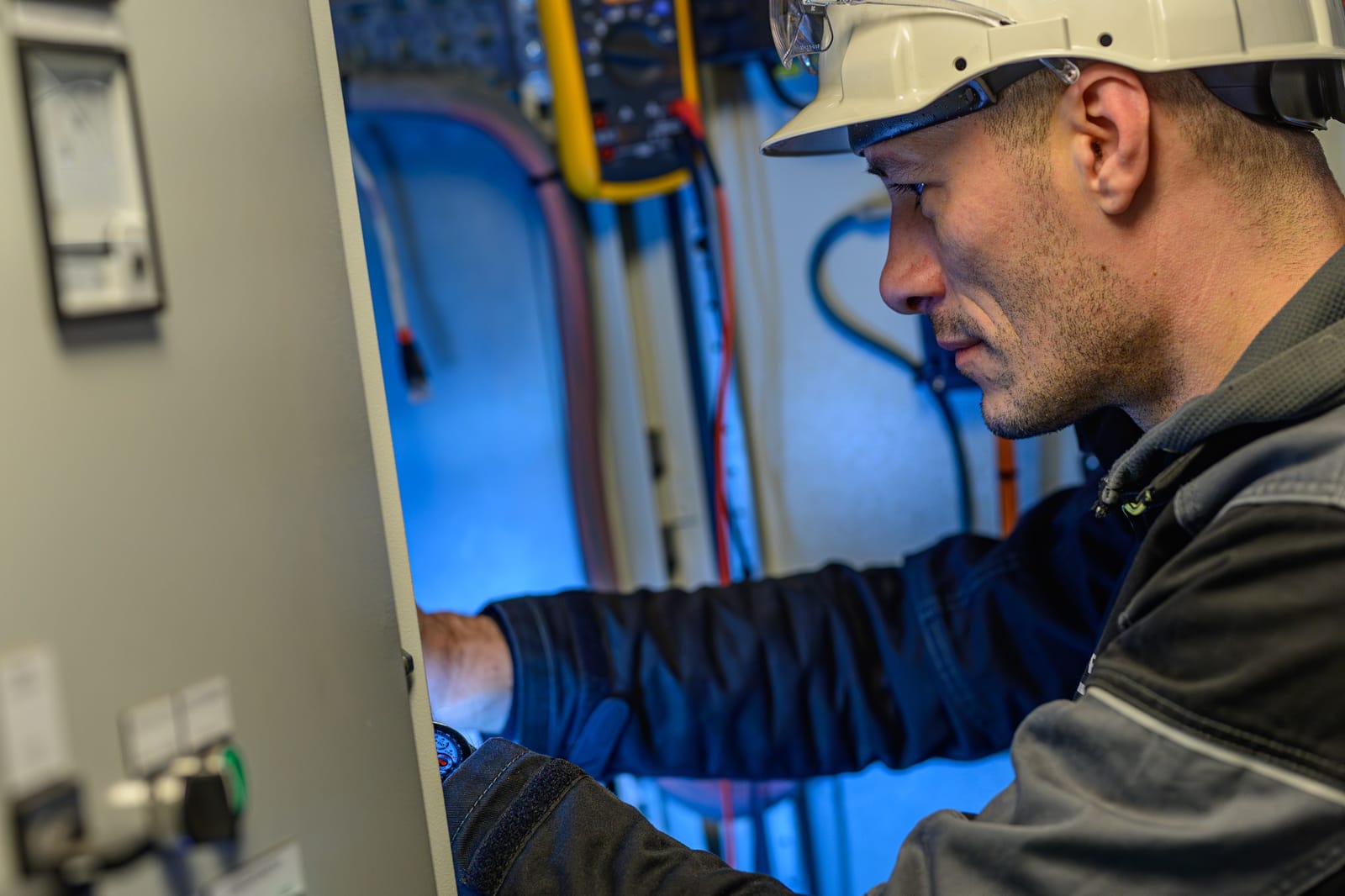 Black Frog Elektro technician in a white hard hat working inside an open marine electrical cabinet, with a multimeter and color-coded wiring visible behind the panel door.
