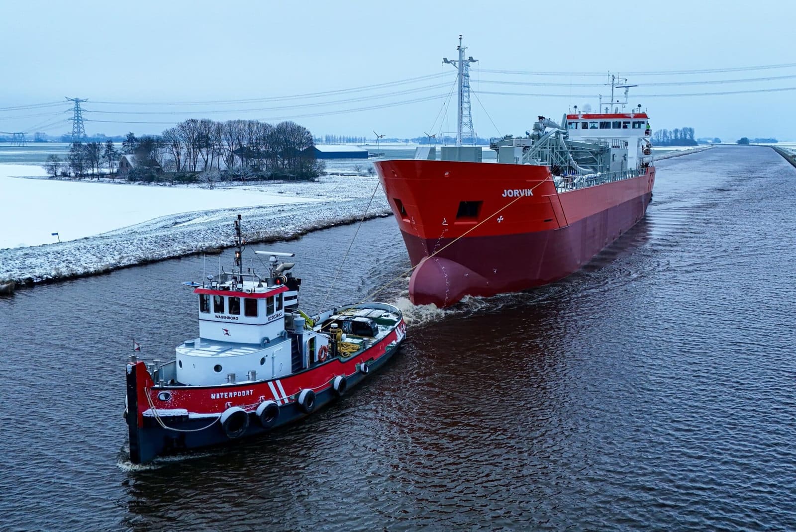 Cement carrier Jorvik for SMT Shipping assisted by tug Waterpoort along a snowy Dutch canal, red hull.