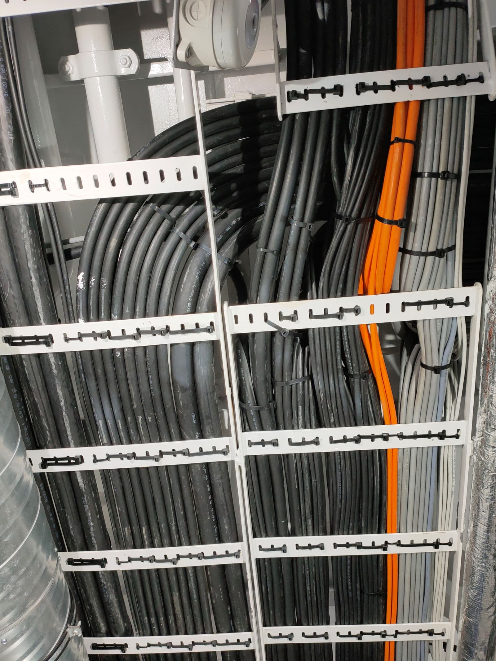 Closely packed grey and orange marine cables on a stacked vertical cable tray rack inside an electrical compartment