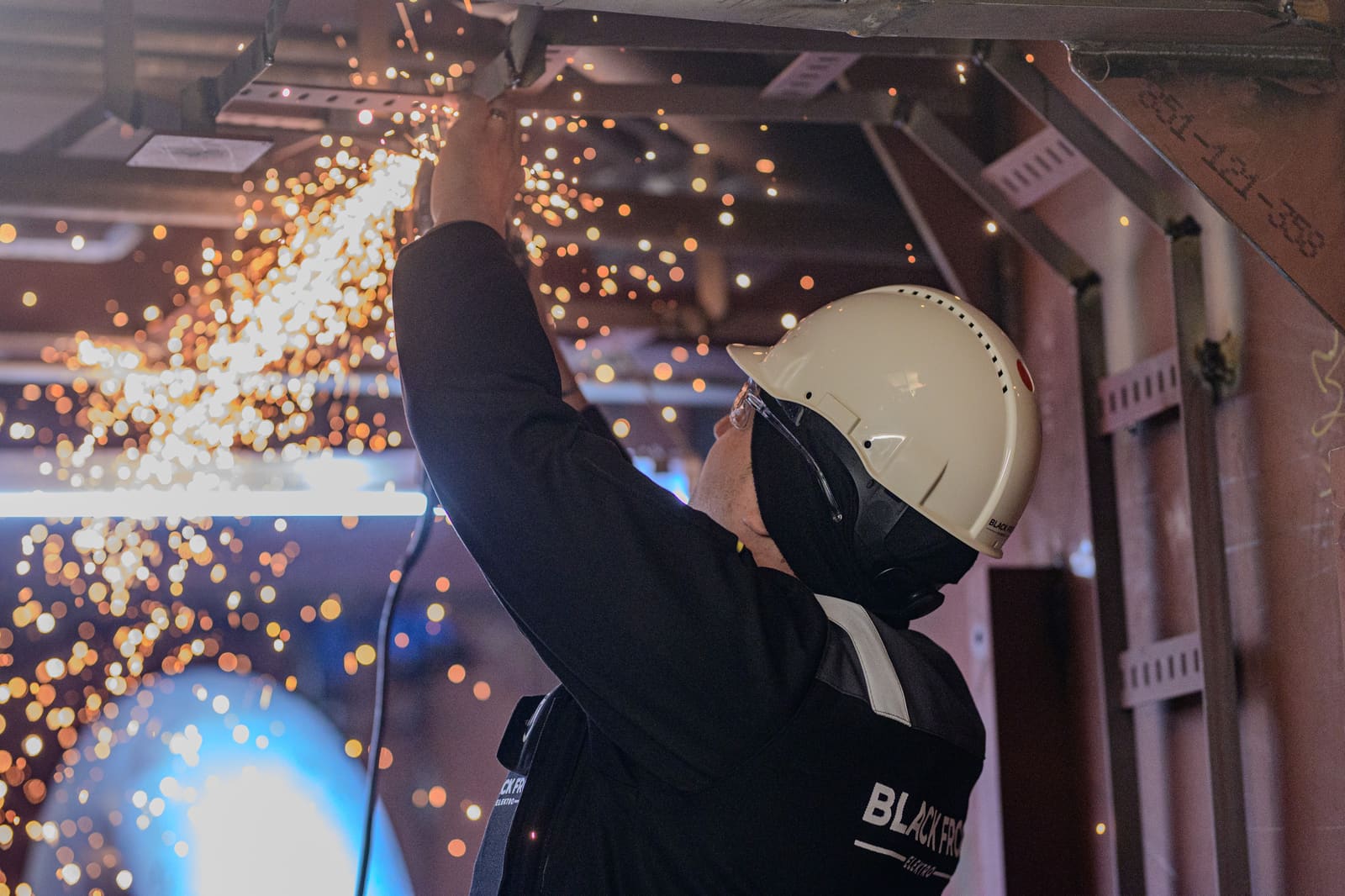 Welder with full-face helmet welding overhead steel cable trays inside a ship compartment, sparks falling down