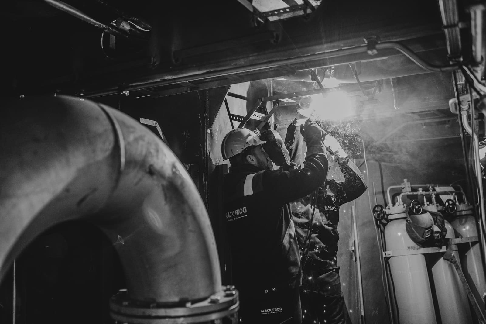 Black-and-white photo of a Black Frog Elektro technician welding overhead in a ship machinery space with industrial piping