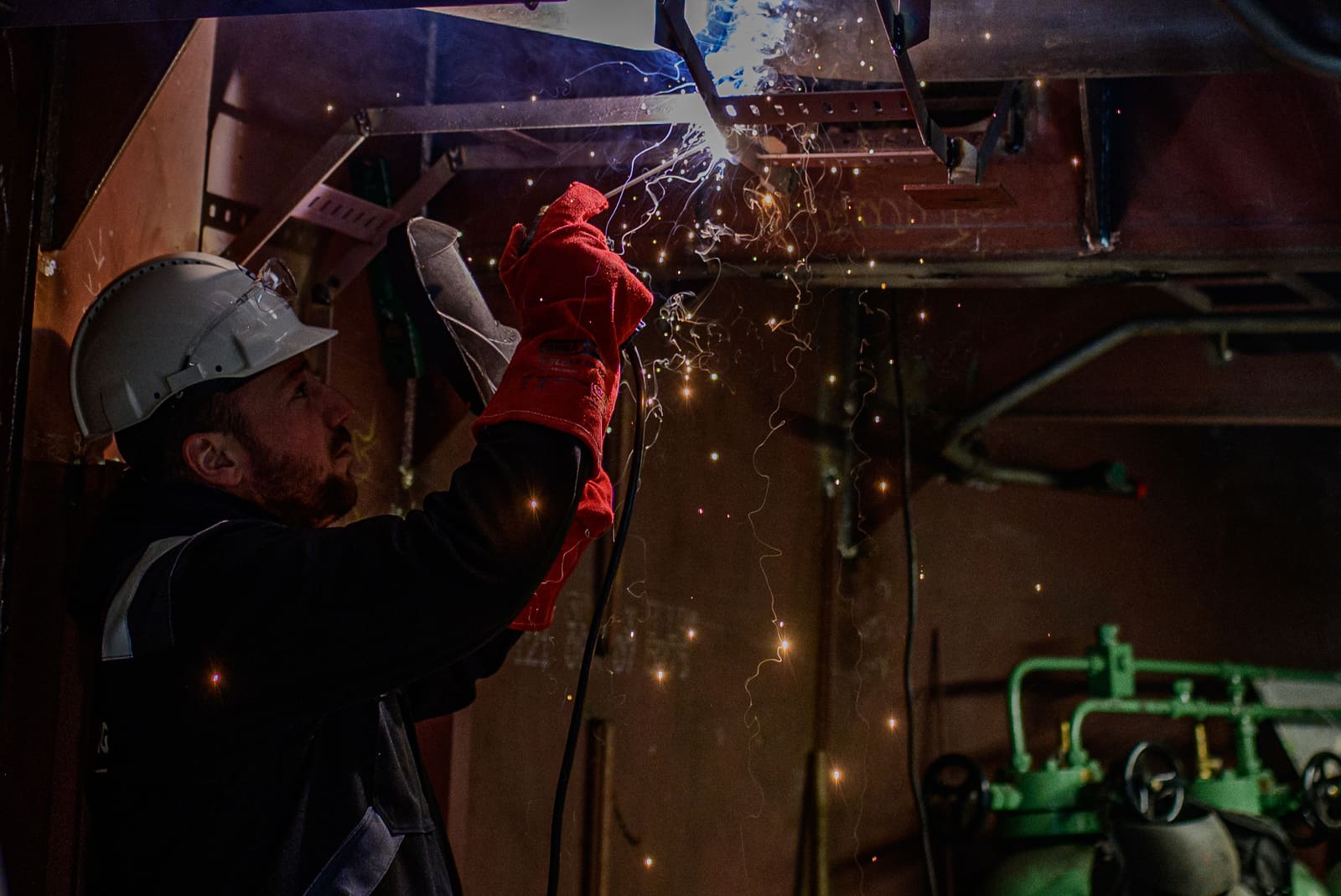 Welder in red safety gloves welding overhead steel structures inside a vessel under construction, sparks raining down